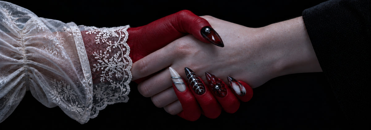A red hand with black and red stiletto press-on nails shaking a hand in a white lace cuff
